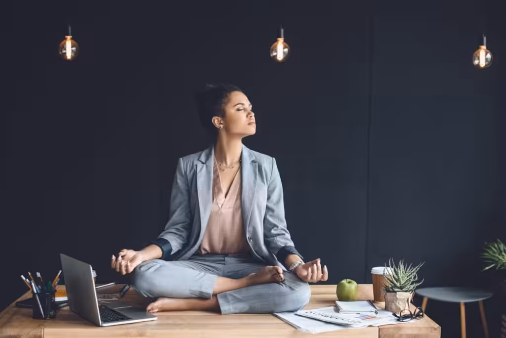 A woman in professional attire sitting on a desk in a meditative pose, illustrating the Mindfulness Based Emotional Regulation in Houston Willowbrook for focus and stress management in a workplace setting.