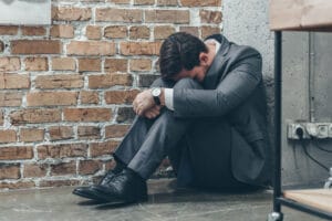 sad man in gray suit sitting on floor and hugging knees on brown textured background in room Acceptance Path Counseling A man in a grey business suit sits huddled on the floor against a brick wall with his head bowed to his knees his posture conveying a sense of profound distress and isolation