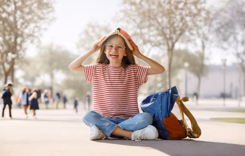 A happy young girl sitting outside with a backpack, playfully balancing a book on her head to represent creative and engaging ways on how to help a child with ADHD focus in school.