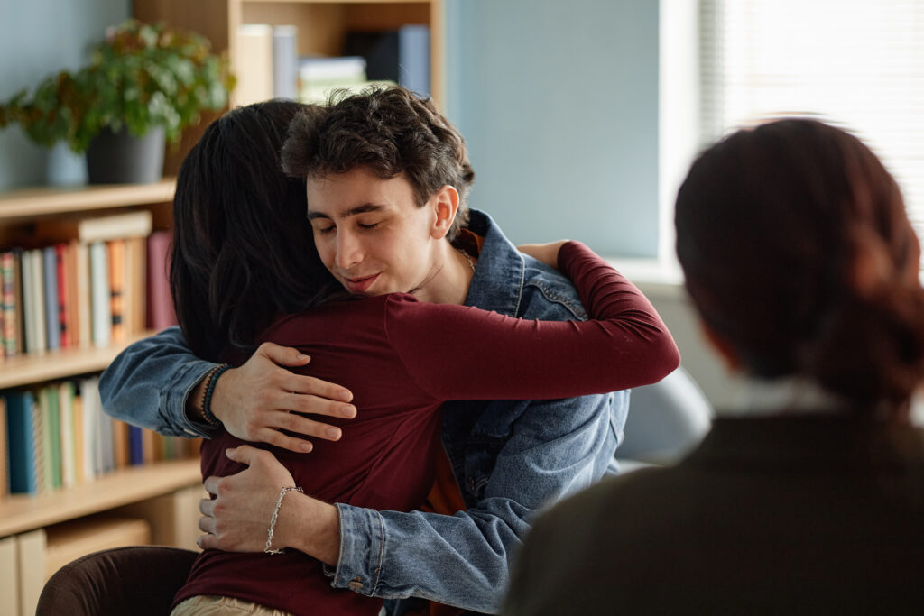 A young man receiving a warm, reassuring embrace from a loved one inside an office or living room space, highlighting the emotional relief found through intentional depression support.