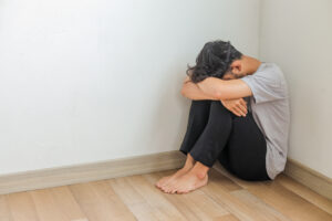 A person curled up tightly on the floor in a corner with their head resting on their knees visually representing the severe social withdrawal and helplessness common to signs of depression