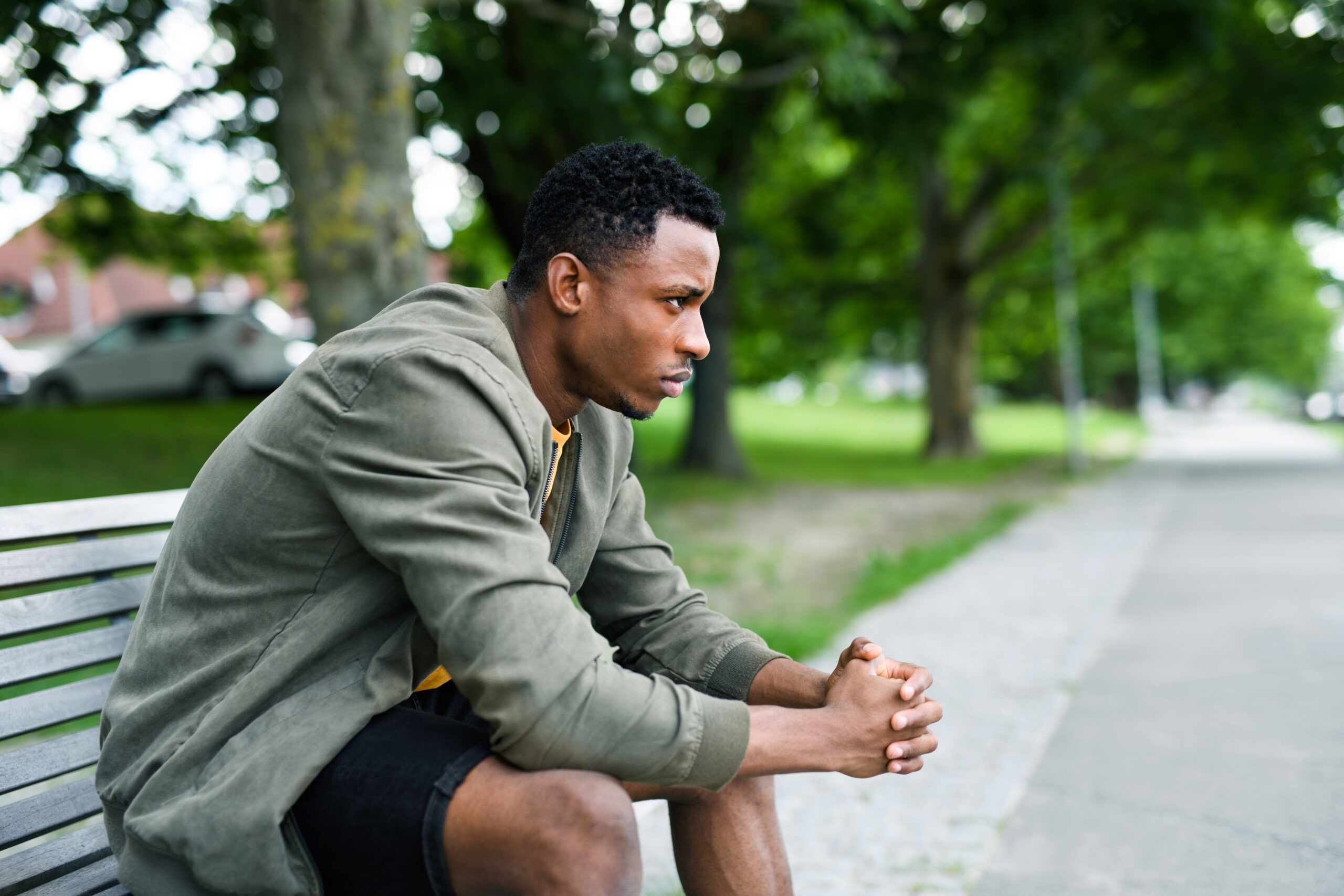 A young black man sits on a park bench looking down with his hands clasped and a serious expression It visualizes the lethargy and social withdrawal common in the initial stages of how to cope with depression