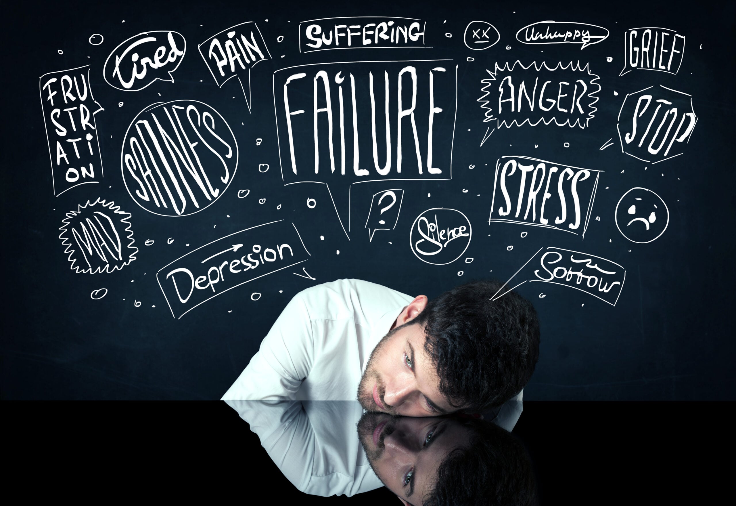A man resting his head on a black surface beneath a chalkboard covered in chaotic words like failure and stress illustrating the overwhelming mental burden and negative thought loops that mark signs of depression