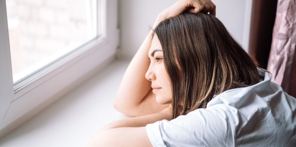A woman looks thoughtfully out a window while resting her chin on her arms. The scene captures a quiet moment of reflection, introducing practical coping mechanisms for depression available to local residents in The Woodlands