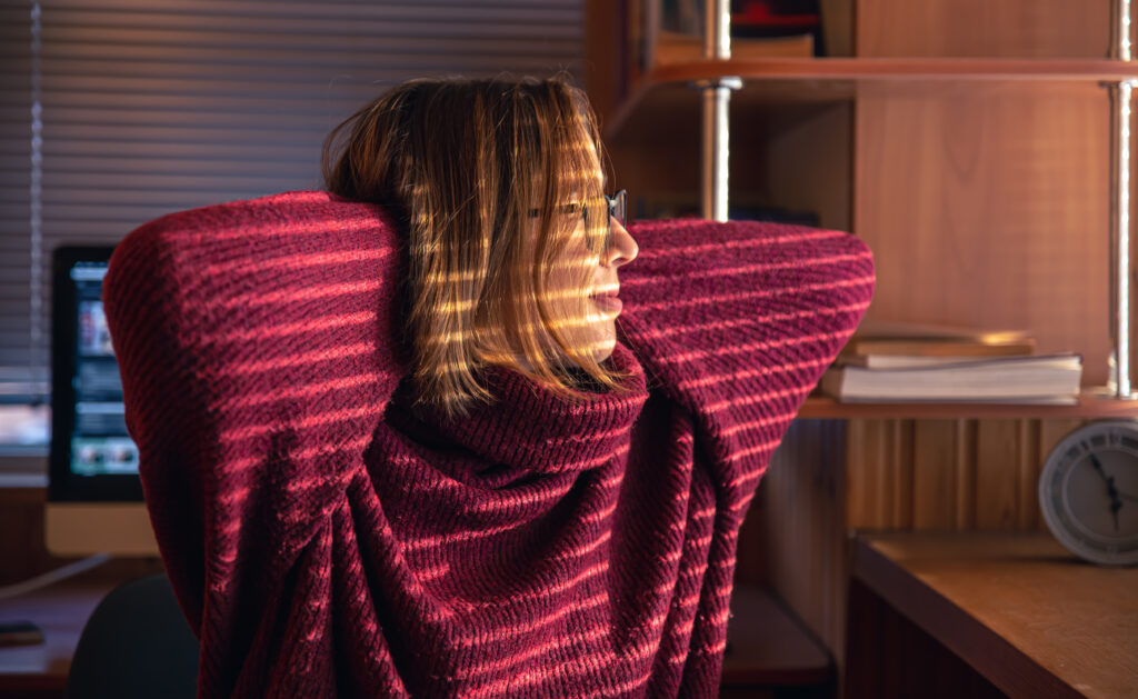 A woman sits in a dark room, looking tensely out through window blinds. The moment captures the constant vigilance and fear common in contamination phobia.