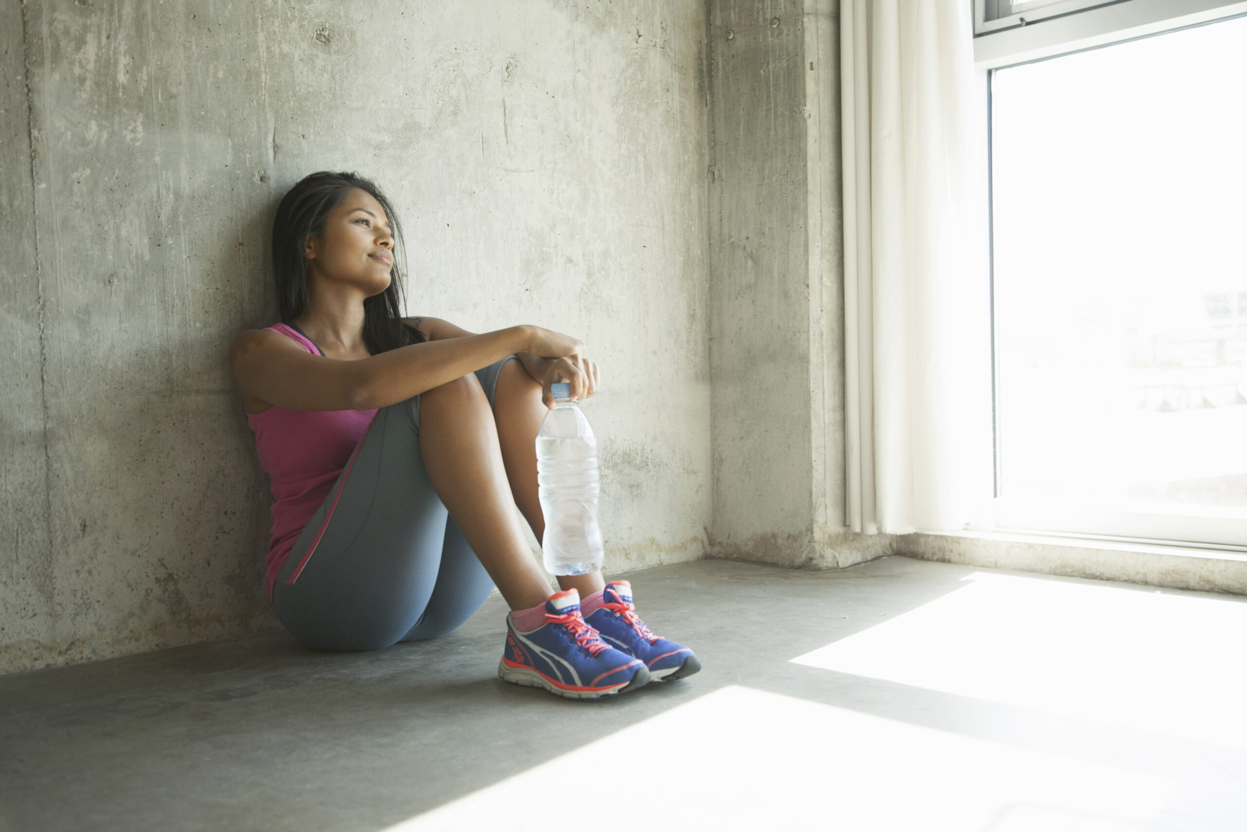 A woman sits on the floor with her back against a wall holding a water bottle after a workout with her eyes closed It captures the extreme physical fatigue and motivational challenges patients experience when learning how to cope with depression