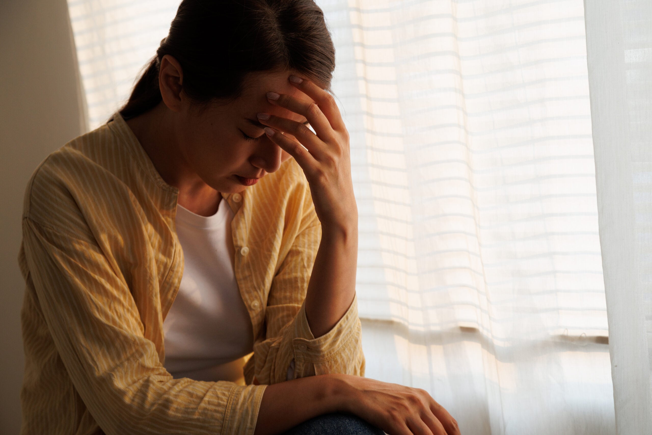 A woman sitting indoors by a window with her hand covering her face in distress capturing the isolation and profound sadness often seen as signs of depression