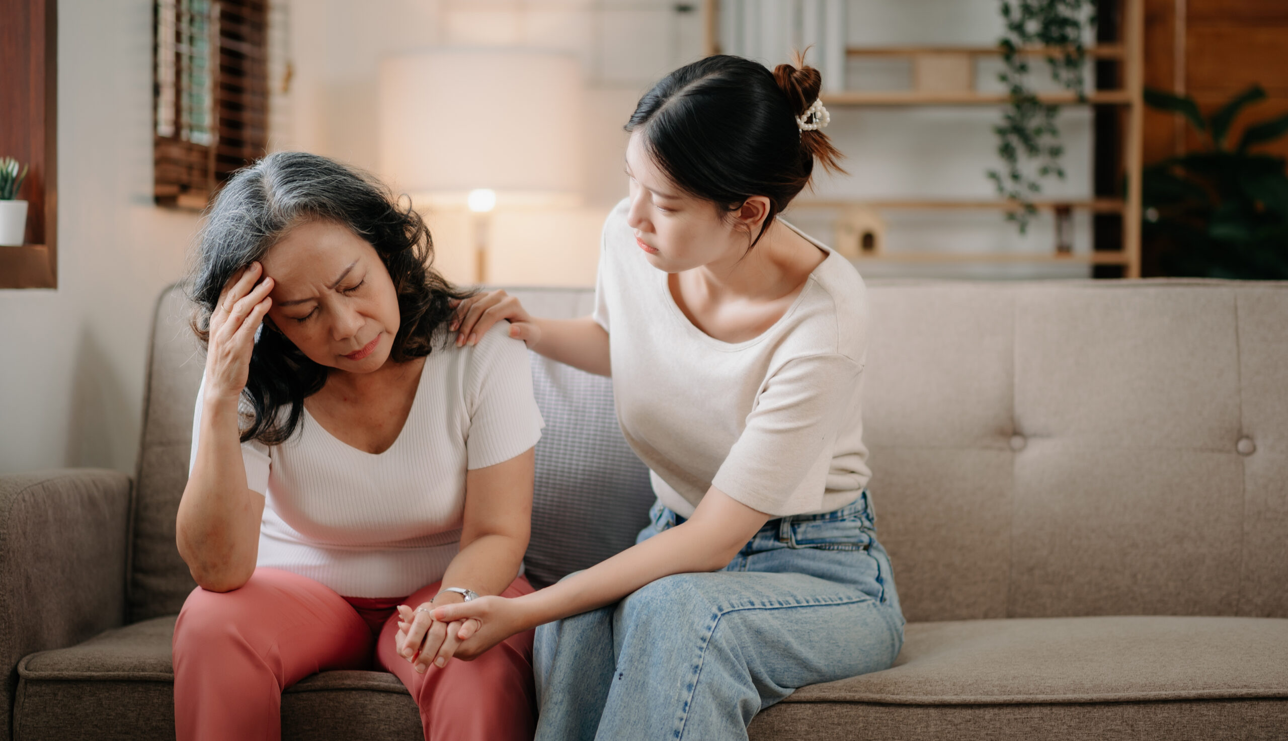 A young woman gently placing a hand on an older woman's shoulder to offer comfort on a living room sofa, highlighting quiet patience when learning how to support someone with depression.