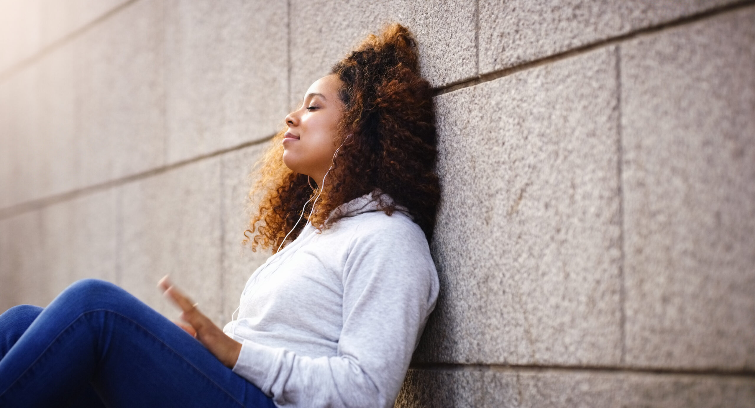 A young woman sitting outside against a textured stone wall with her eyes closed practicing mindful breathing exercises to focus on overcoming depression