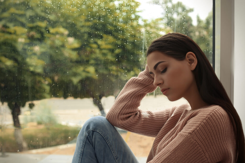 A woman in a pink sweater sits by a rainy window with her eyes closed, leaning her head against her hand. The image captures a quiet moment of reflection, introducing supportive strategies on how to cope with depression