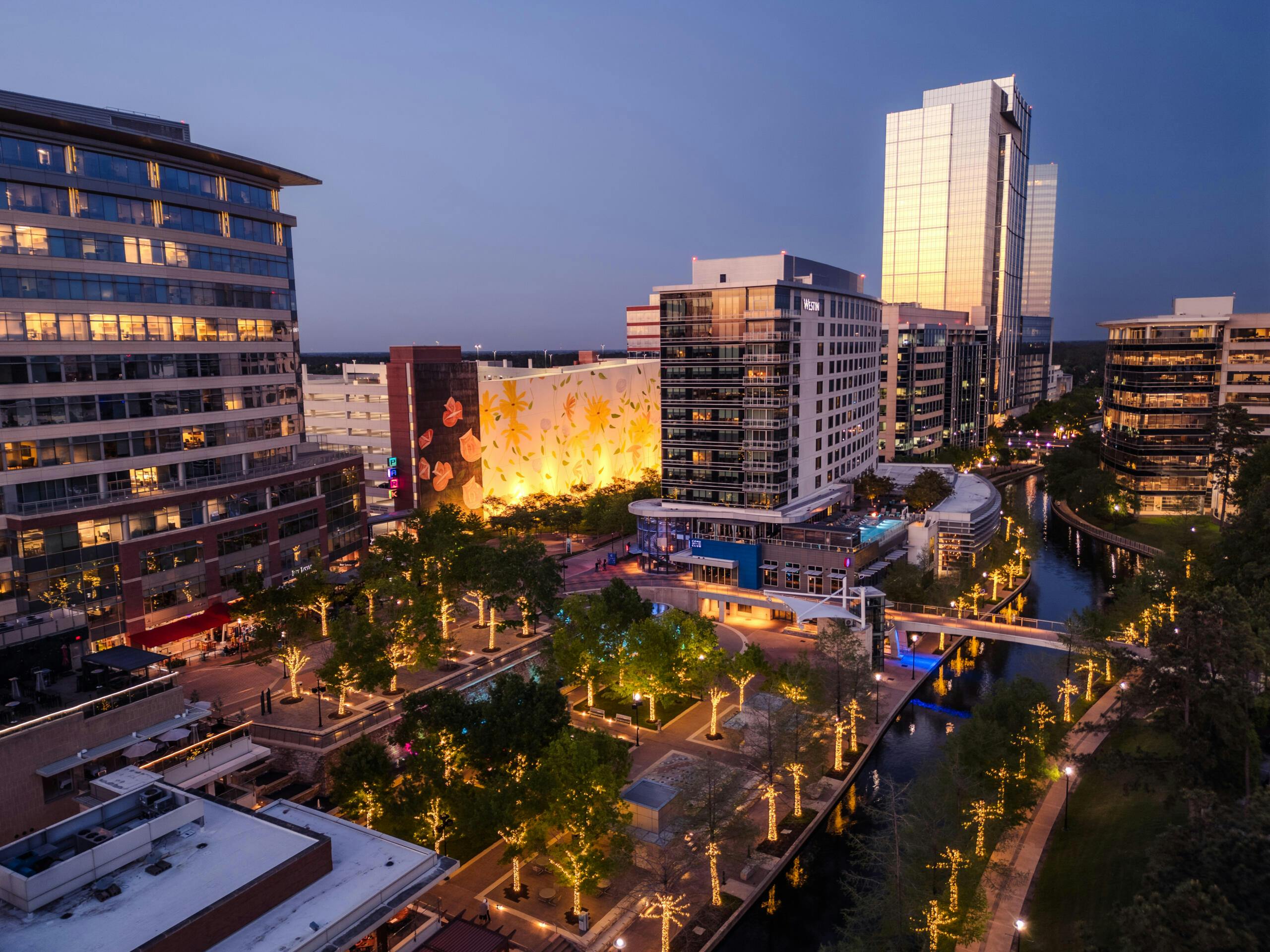 Aerial view of a modern downtown at dusk with glass office buildings and a lit waterfront promenade along a canal