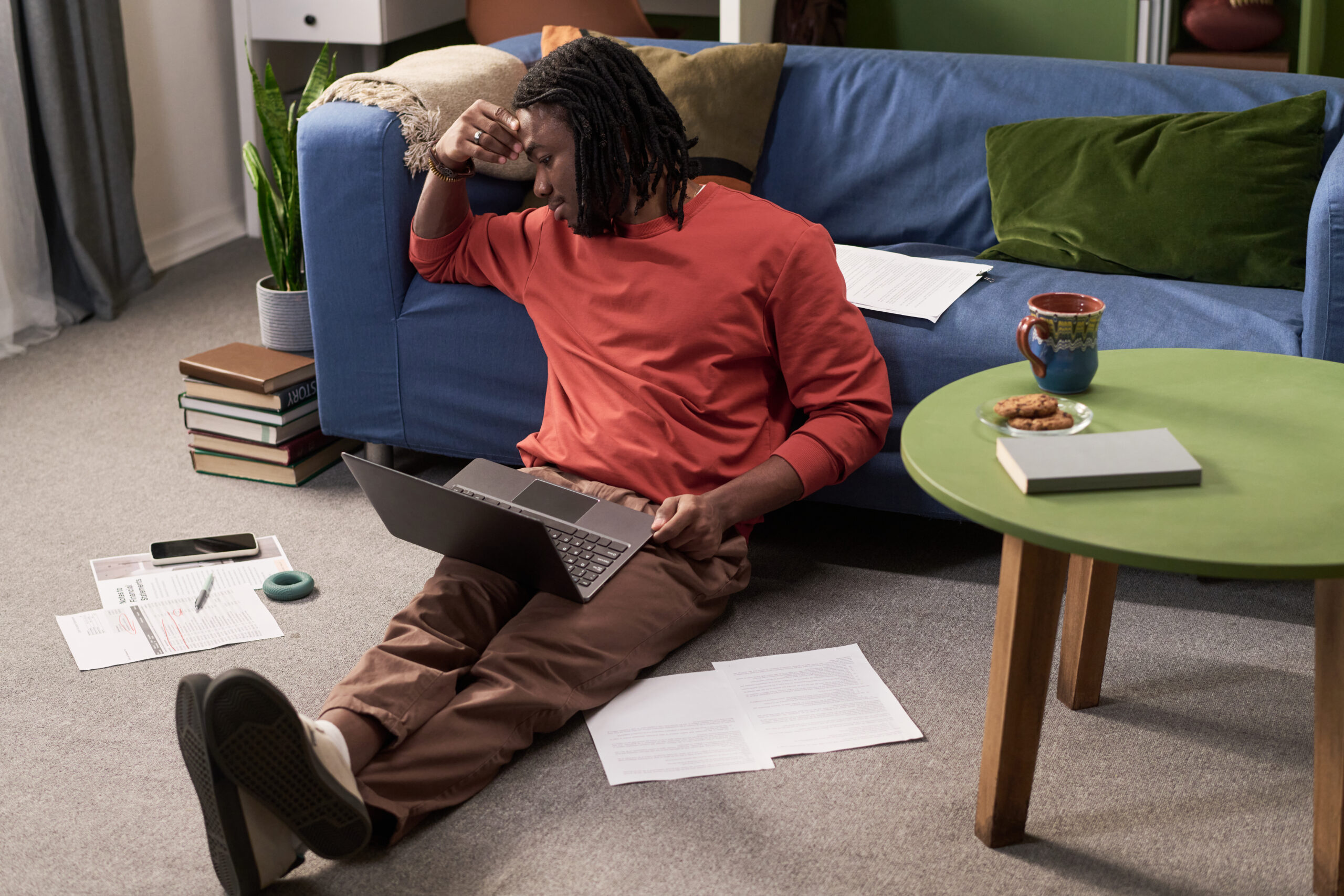 Young Black Man Sitting on Floor Studying with Laptop Looking Stressed Acceptance Path Counseling Overwhelmed person looking at an unfinished task list and scattered notes