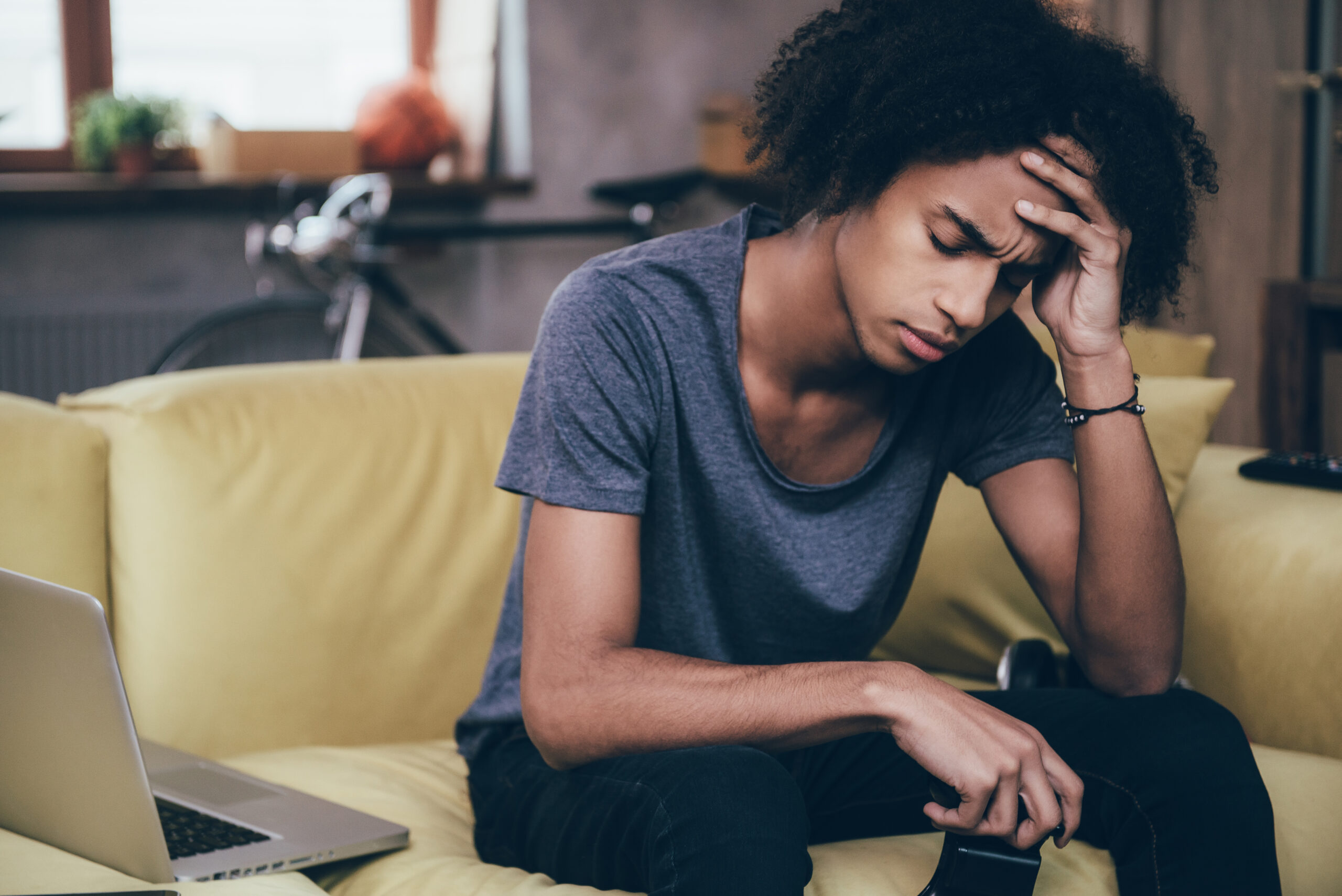 Lost that battle Desperate young African man holding joystick and keeping his hand on forehead while sitting on the couch at home Acceptance Path Counseling Unfinished task list illustrating the cycle of depression and procrastination