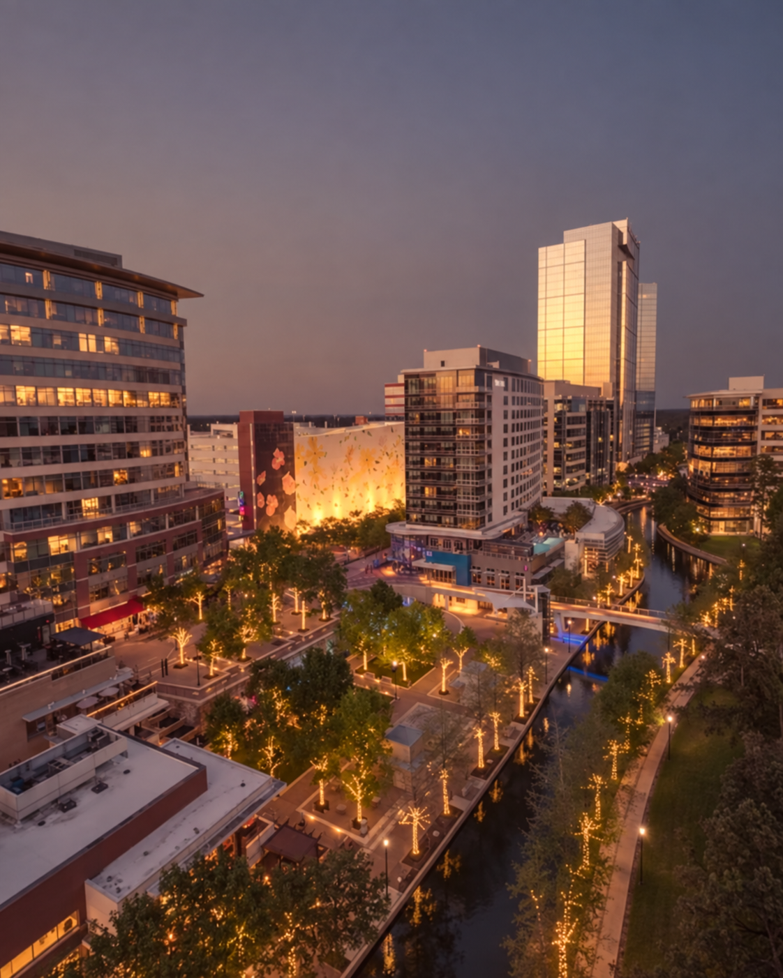 Aerial view of a modern city plaza at dusk with illuminated trees lining a canal and tall glass buildings in the background