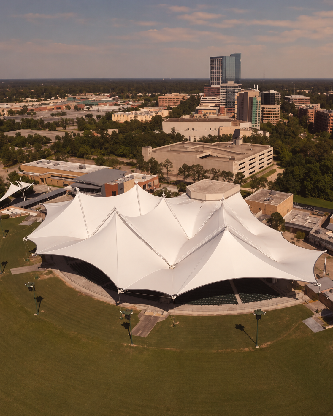 Aerial view of a large white tent like canopy over an outdoor amphitheater in a park with city buildings in the background