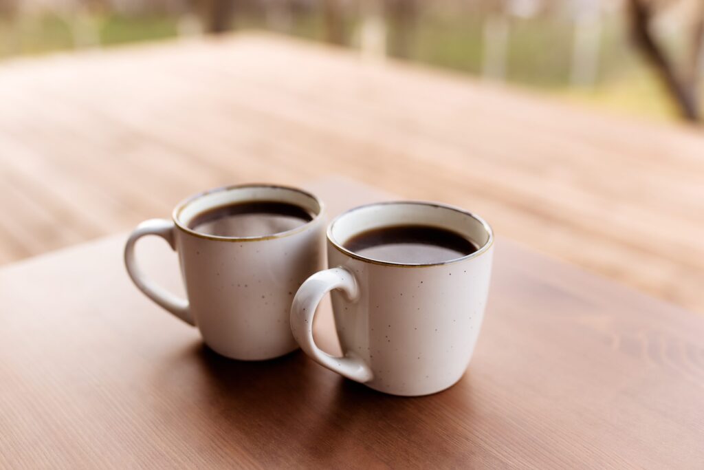 Two white mugs of black coffee sit close together on a wooden table