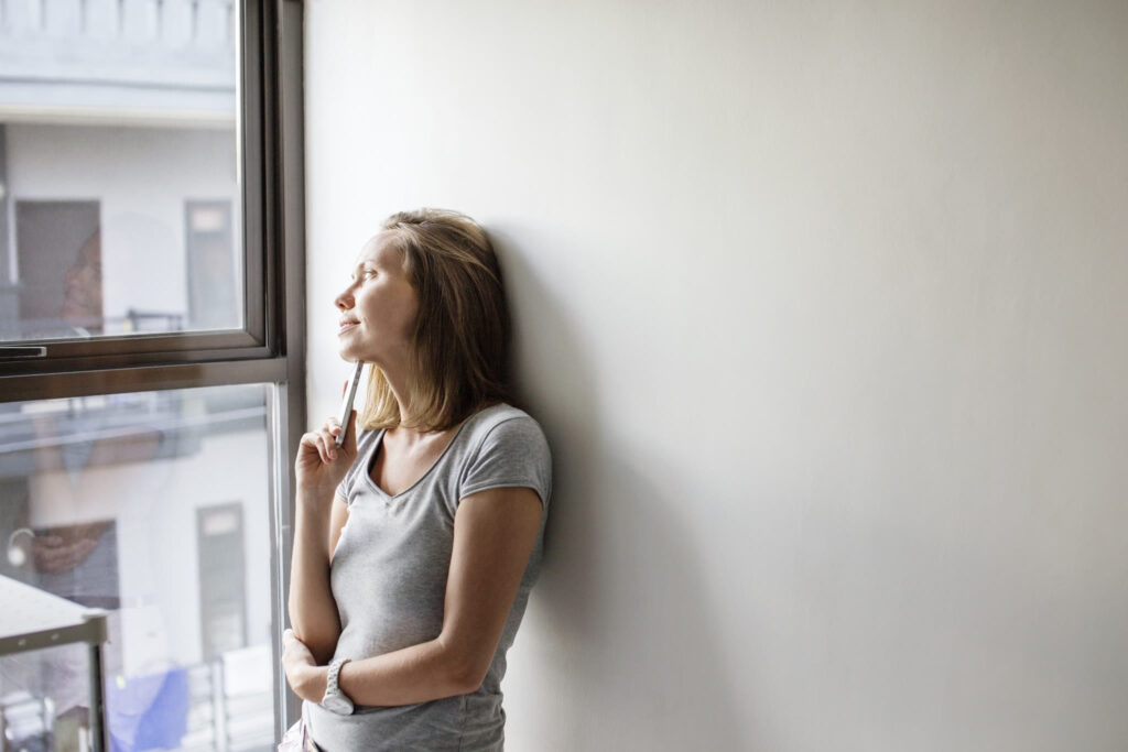 A woman in a gray top leans against a white wall and looks out a window